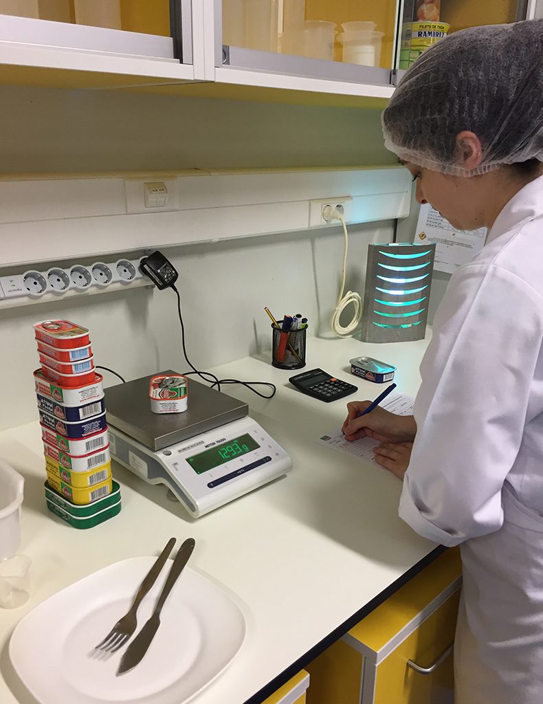 Técnica do laboratório Ramirez confere na balança o peso de uma lata de sardinha, com outras latas empilhadas ao lado